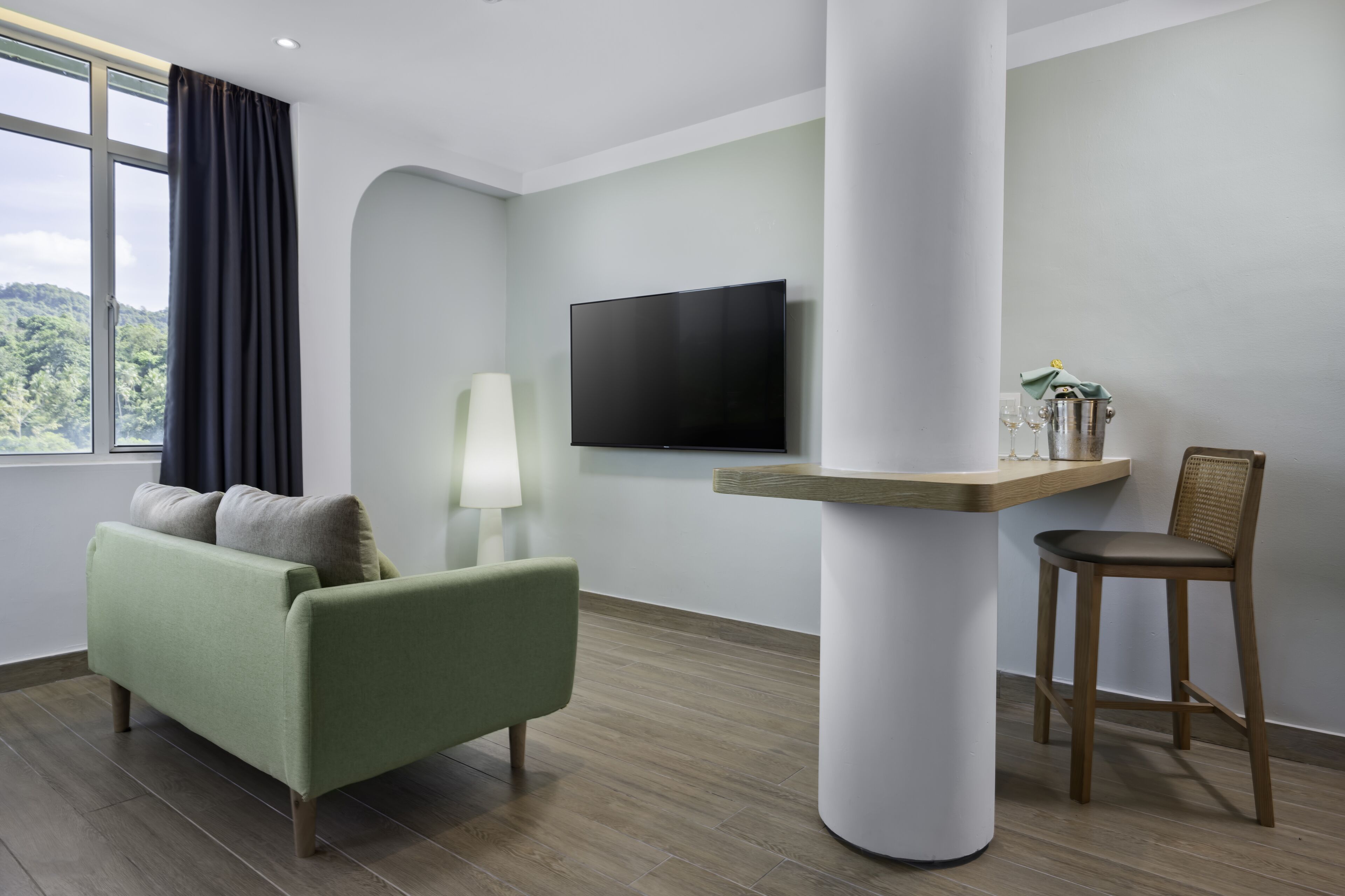 A modern room with a green sofa, a wooden chair, a flat-screen TV, and a table with a lamp and a bucket.