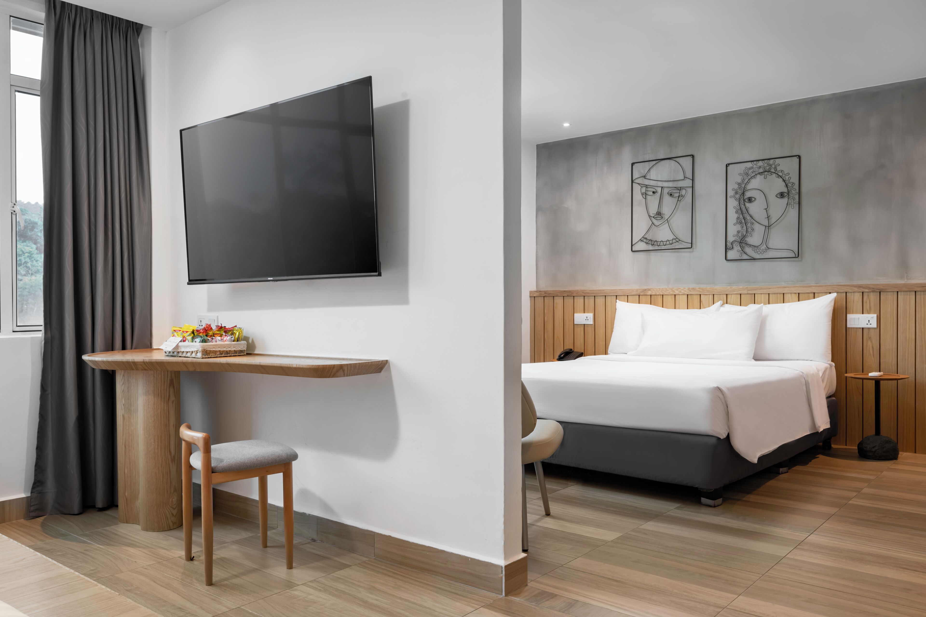 Une chambre d’hôtel avec un lit, un téléviseur fixé au mur, une table en bois avec une chaise et des œuvres d’art accrochées au mur.