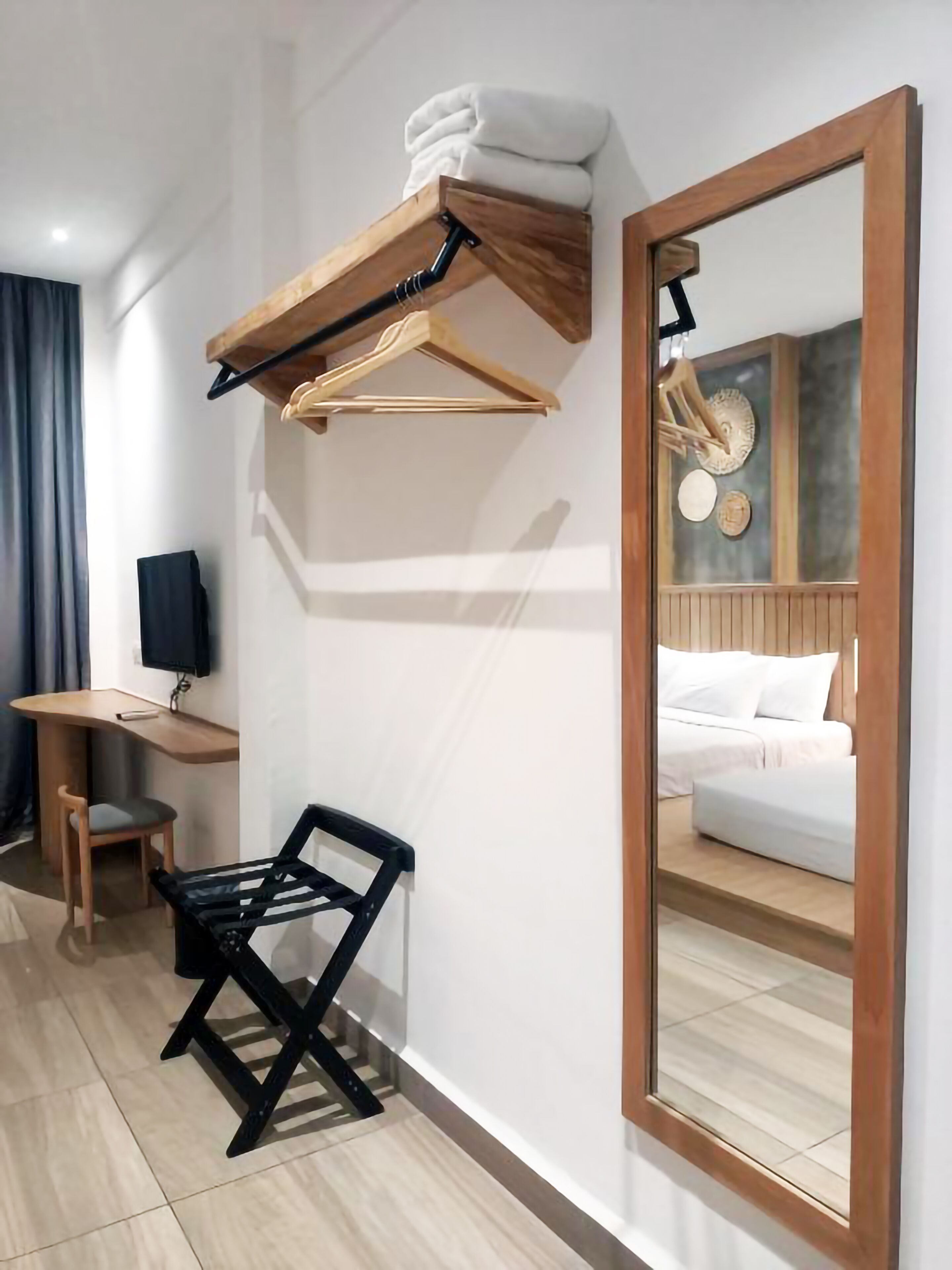 A hotel room with a wooden desk, a black chair, a hanging wooden rack with towels, a large mirror, and a bed with white bedding.