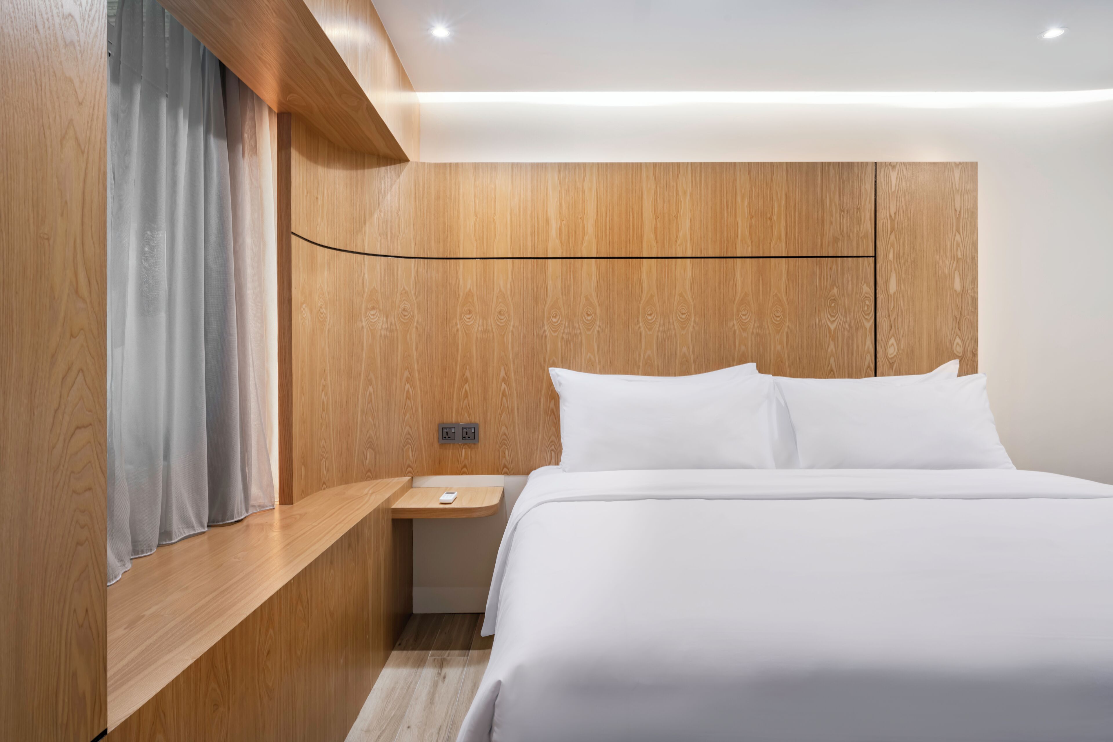 A modern hotel room with a wooden headboard, a bedside table with a lamp, and a large bed with white linens.