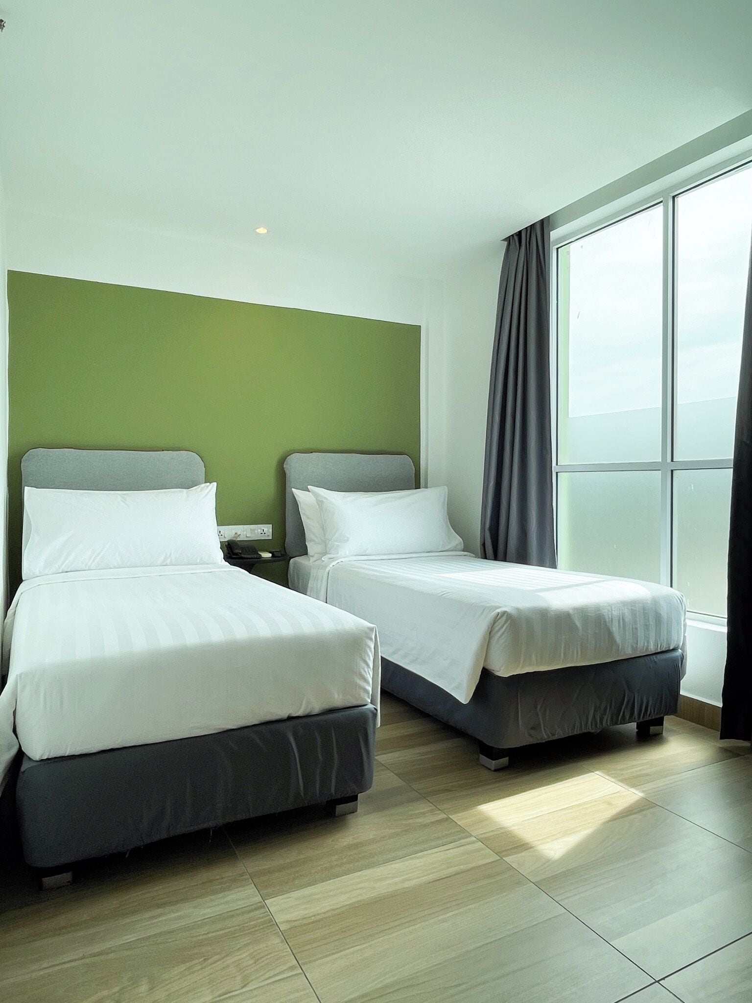 A hotel room with two beds, a green accent wall, and a large window with curtains.