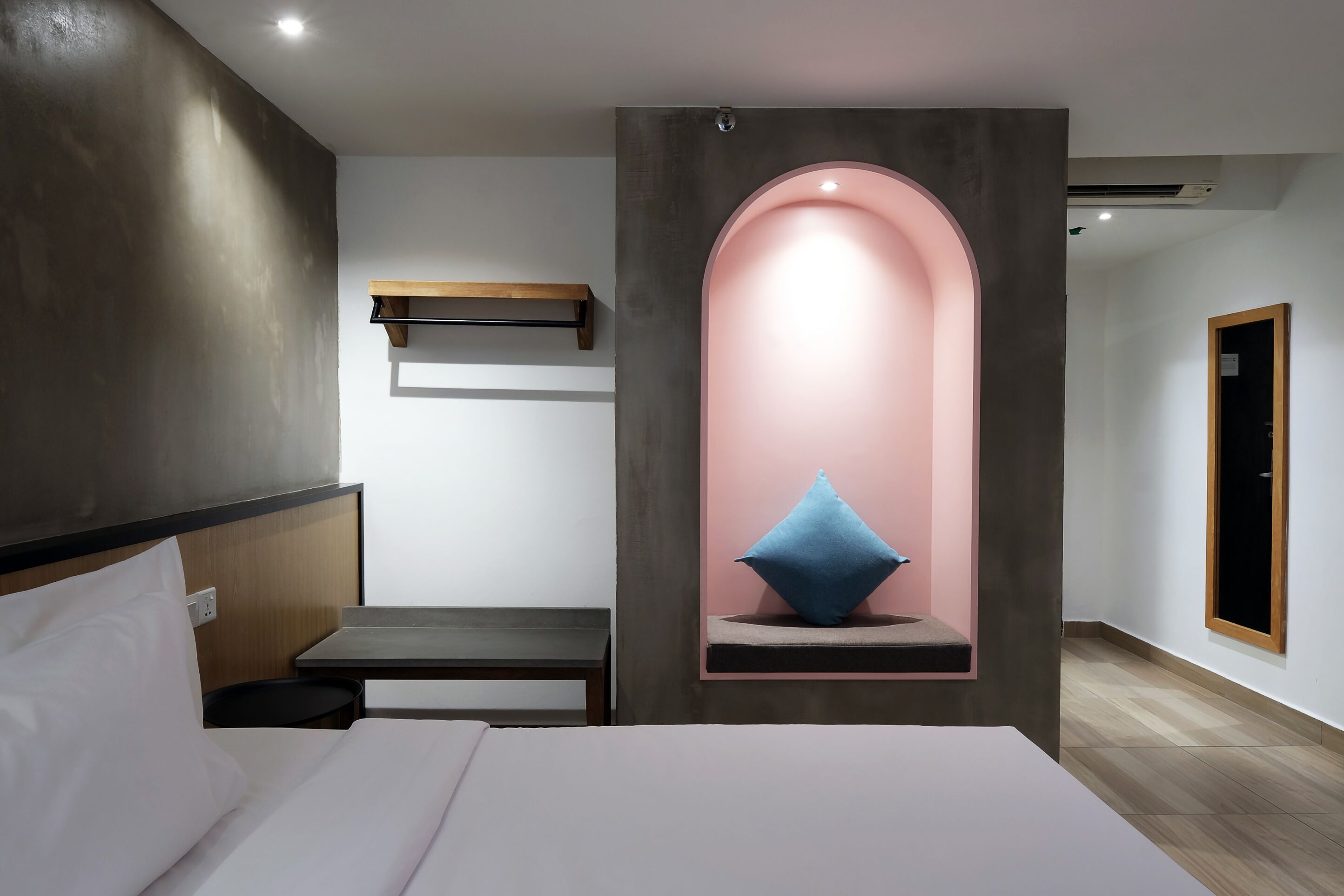 A modern hotel room with a bed, bedside table, and a unique pink archway with a blue pillow.