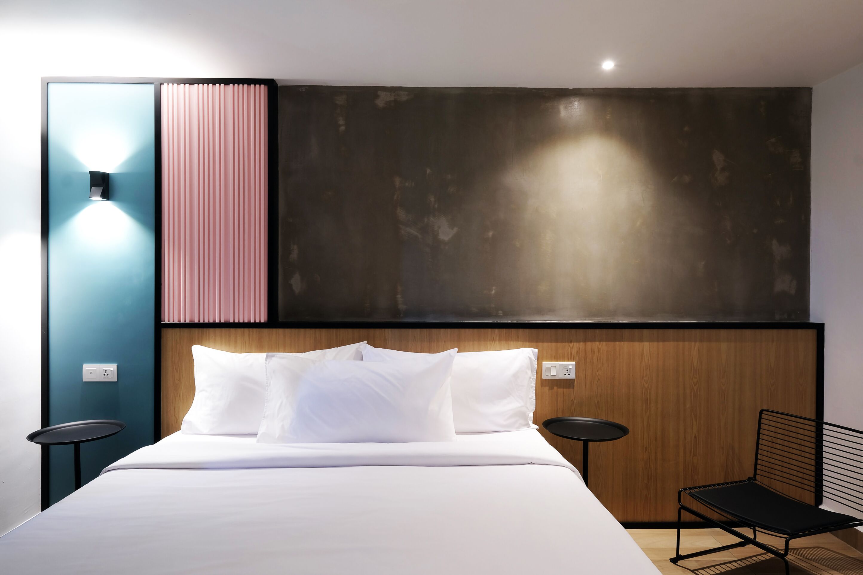 A modern hotel room with a large bed, a wooden headboard, a small round table, and a black metal chair.