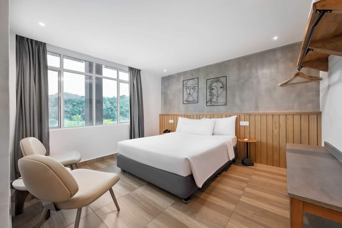 Une chambre d’hôtel moderne avec un grand lit, un bureau et une chaise. Elle comprend deux fenêtres avec des rideaux, une table de chevet avec une lampe et des œuvres d’art accrochées au mur.