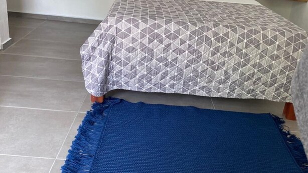 A bed with a geometric patterned bedspread, a blue woven rug, a macramé wall hanging, and a small bedside table with a lamp. - Casa Baloo (El Cuyo)