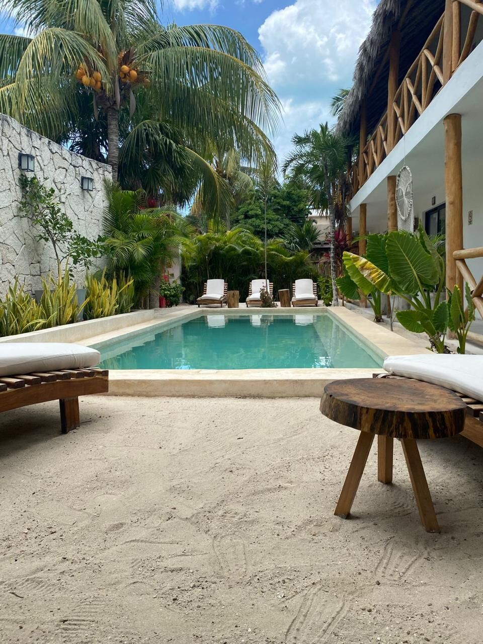Ein Poolbereich mit Liegestühlen, einem Holztisch und tropischen Pflanzen.