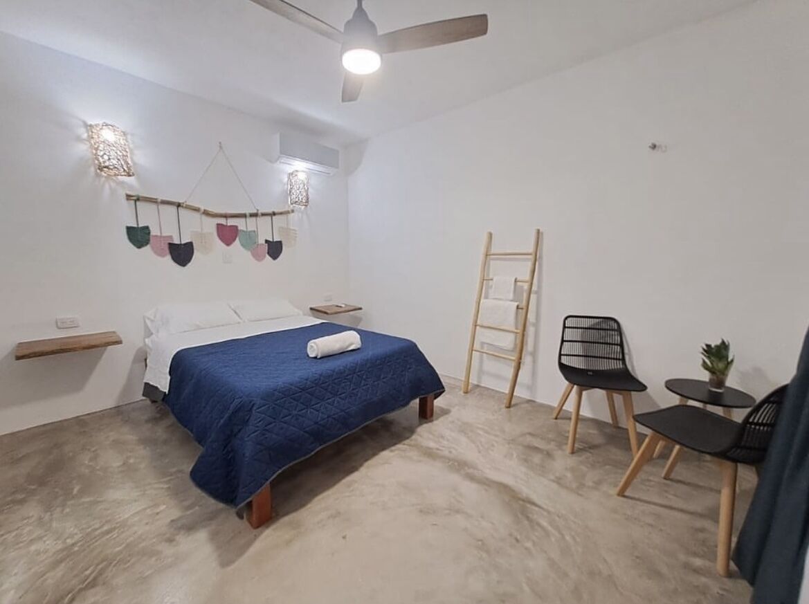 Un dormitorio con cama, ventilador de techo, una mesita con planta y una silla.