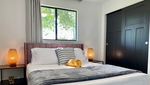 A bedroom with a large bed, two bedside lamps, a window with a view of trees, and a dark door. - Newly Remodeled Beauty in Phoenix (Phoenix)