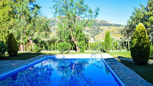 A swimming pool with a stone border, surrounded by greenery and trees, with a mountainous backdrop. - Vacation Home 'El Rancho' with Mountain View, Pool, Wi-Fi & Garden (El Gastor)