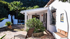 A white building with a covered entrance, a potted plant, a chair, and a stone wall. - Vacation Home 'El Rancho' with Mountain View, Pool, Wi-Fi & Garden (El Gastor)