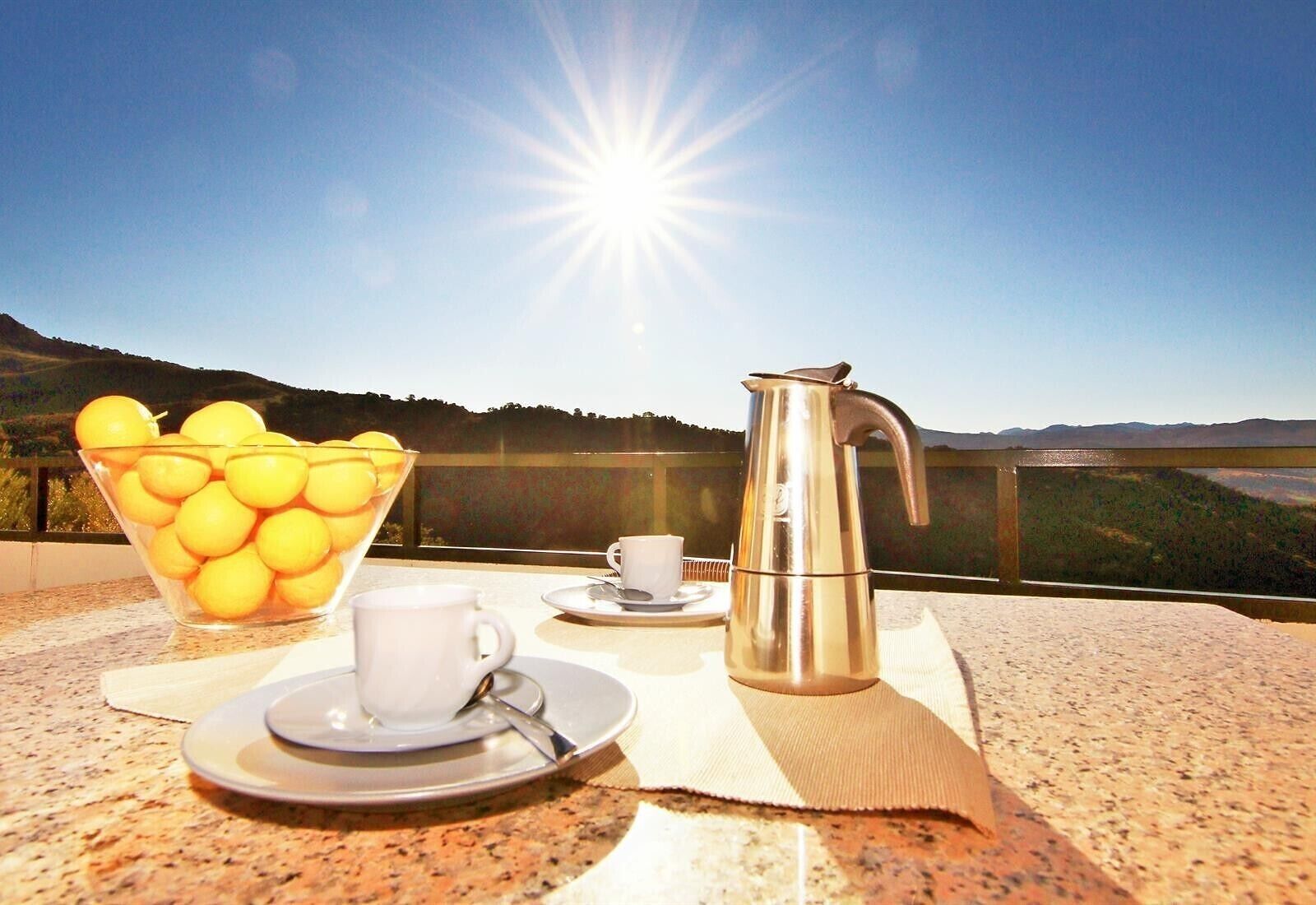 A breakfast setup with a bowl of fruit, a coffee pot, and a cup on a terrace with a scenic view.