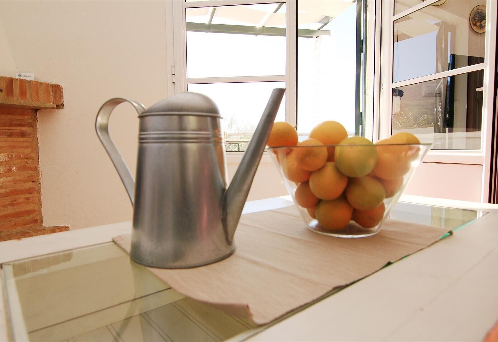 A metal watering can and a clear glass bowl filled with oranges on a table.