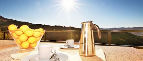 A breakfast setup with a bowl of fruit, a coffee pot, and a cup on a terrace with a scenic view.