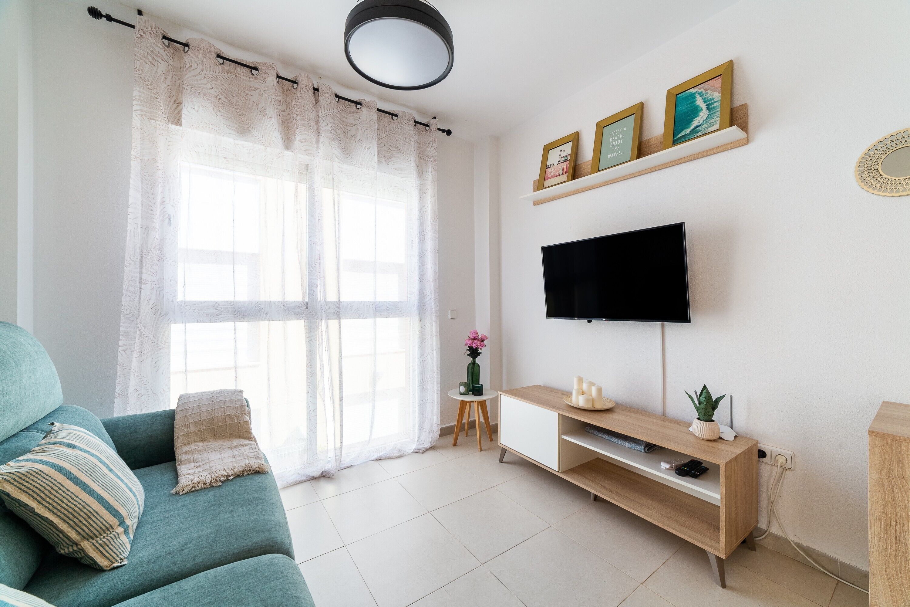 A living room with a teal sofa, a TV, a small side table with a vase, and a window with sheer curtains.