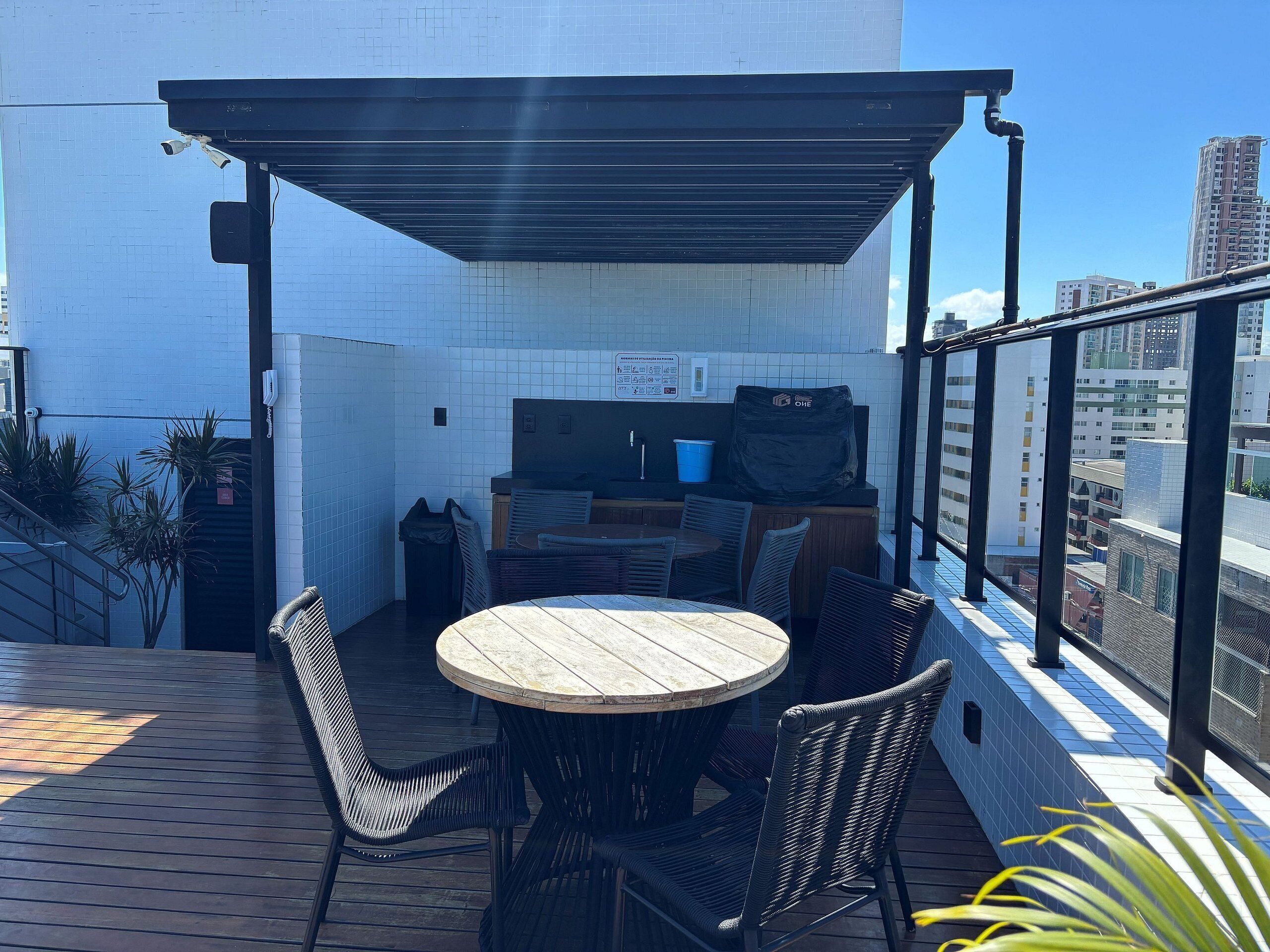 A rooftop patio with a round wooden table, black wicker chairs, a grill, and a view of the cityscape.