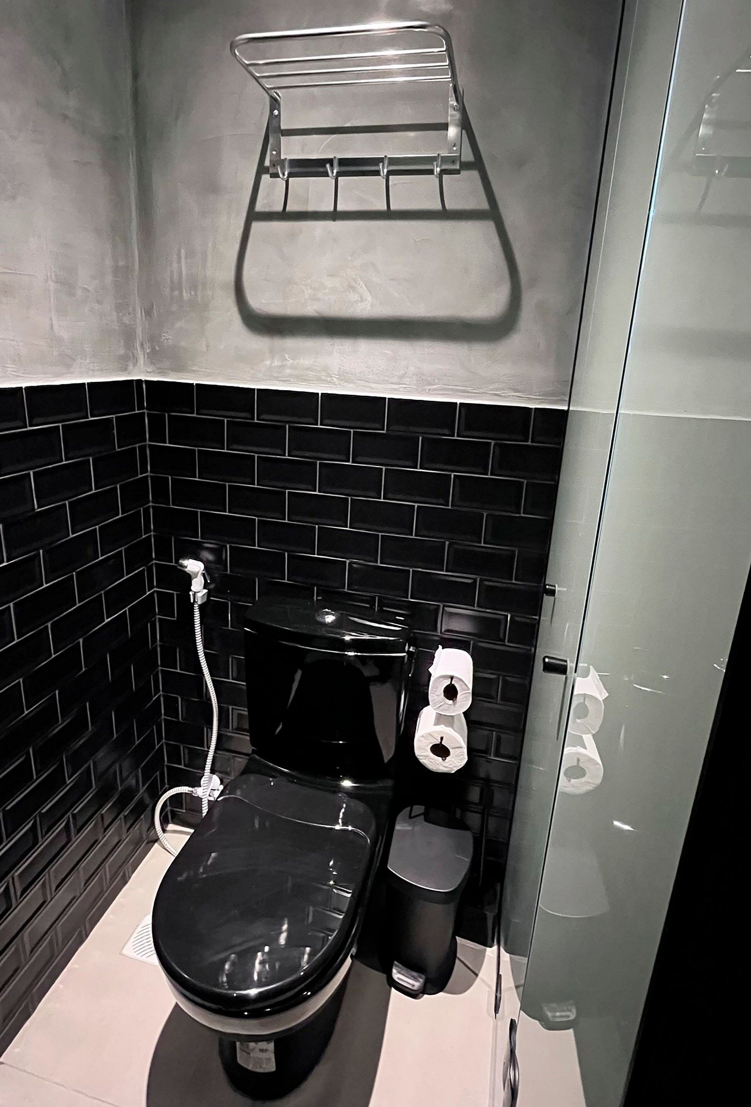 A compact bathroom with a black tiled wall, a black toilet, a white trash bin, and a towel rack on the wall.