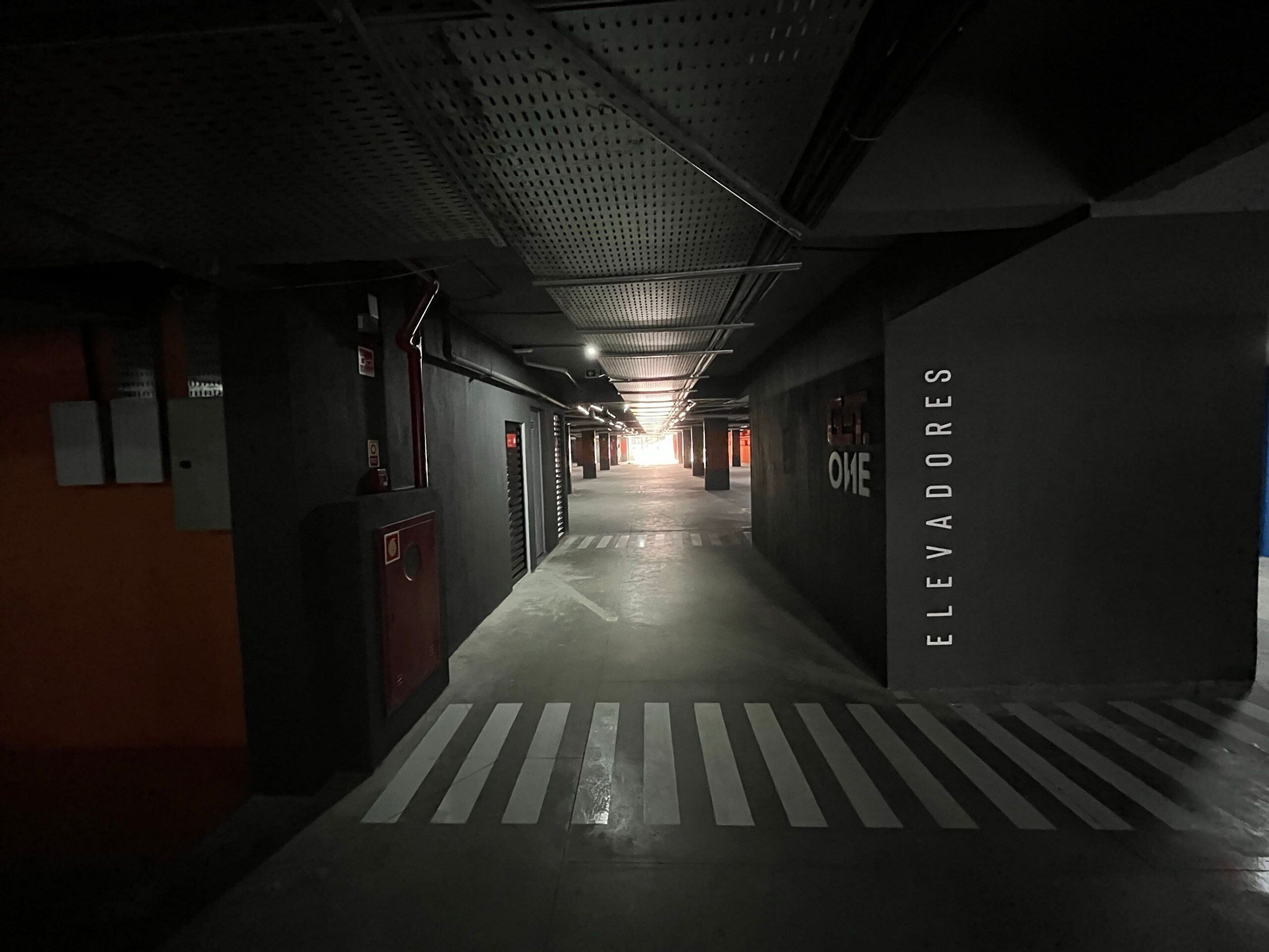 Un couloir faiblement éclairé, avec un sol mouillé, un passage piéton balisé et un mur sur lequel on peut lire le mot « ASCENSEURS ».