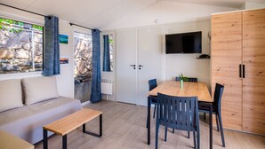 A modern room with a sofa, a wooden table, and chairs, a TV mounted on the wall, and a stone wall with a window. - Mobil-Home Baia with Sea View and Private Terrace (Saint-Raphaël)