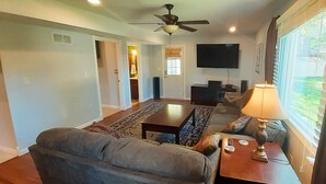 Ein Wohnzimmer mit einer Couch, einem Couchtisch und einem Fernseher. Es gibt einen Deckenventilator, ein Fenster mit Vorhängen und eine Tür zu einem anderen Raum.