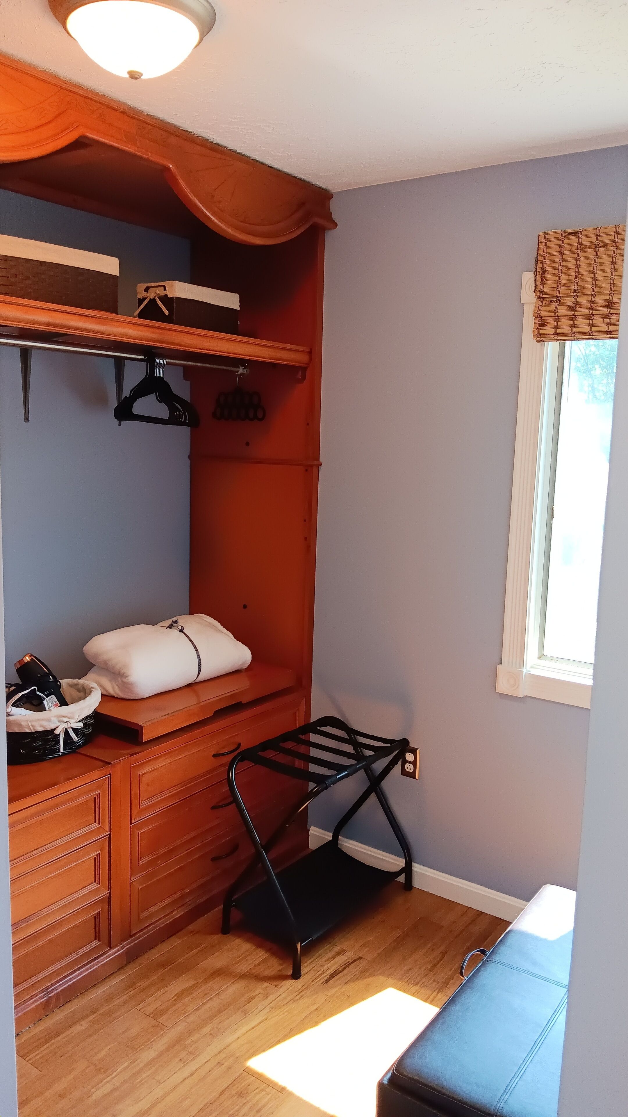A compact bedroom with a wooden dresser, a bed with a white pillow, a black drying rack, and a window with a view of trees.