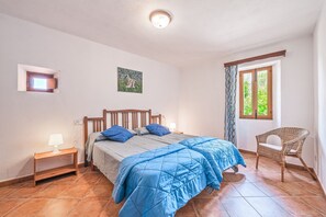 Een slaapkamer met een bed, nachtkastje, lamp, raam, stoel en een schilderij aan de muur.