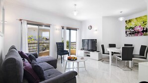 A modern living room with a sofa, dining table, chairs, a TV, and a balcony with a view. - Apartment with Marina and Sea Views – Shared Pool & Wi-Fi (Albufeira)