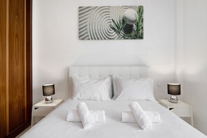 A hotel room with a white bed, two bedside tables, white pillows, a wall art piece, and a wooden door.