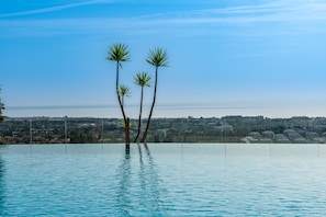 Une piscine avec trois palmiers au centre, entourée d’un ciel bleu clair et surplombant un paysage urbain lointain.
