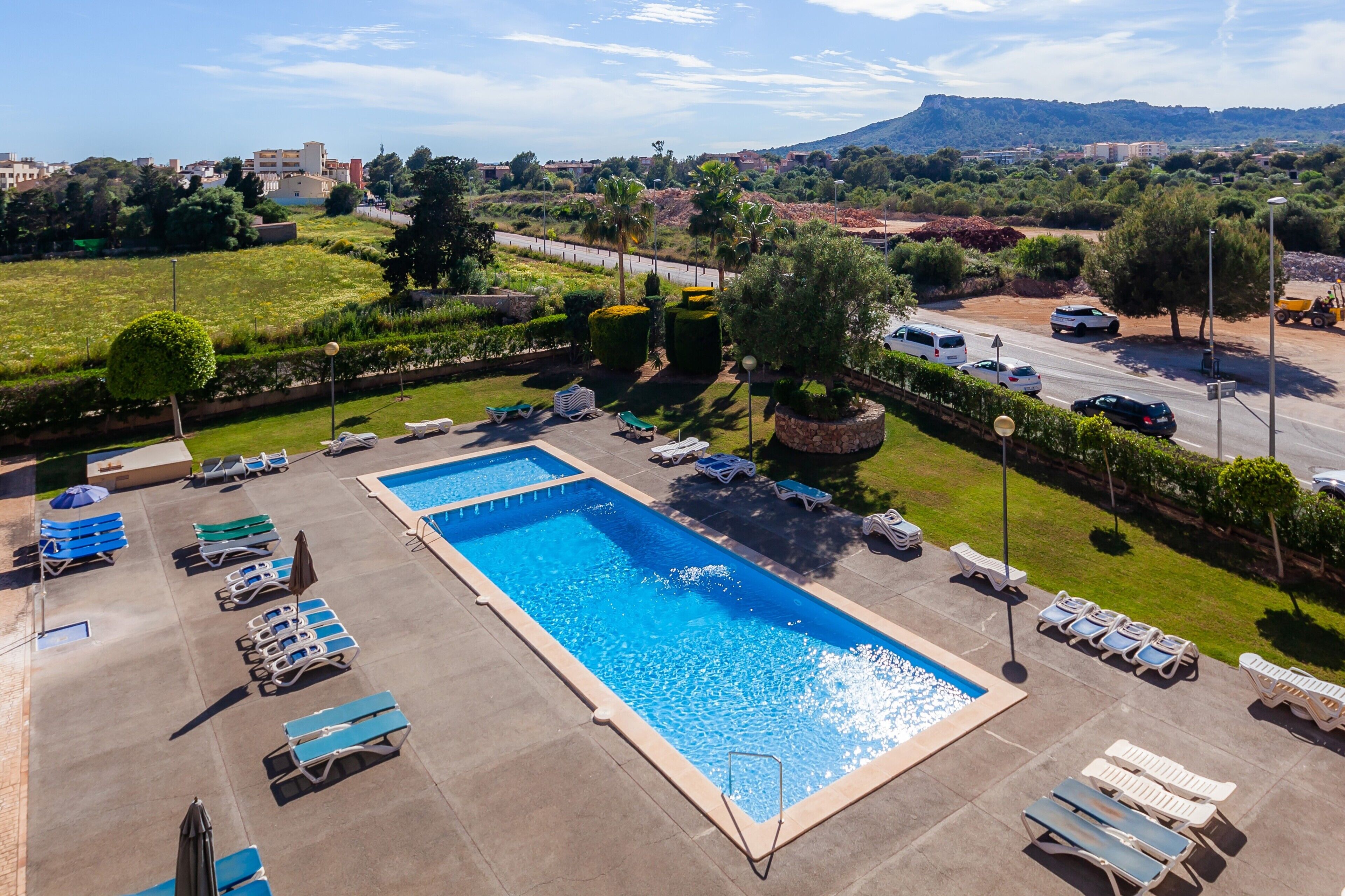 A swimming pool with lounge chairs and umbrellas, surrounded by greenery and a parking area.