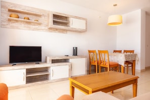 A living room with a TV, wooden cabinets, a dining table with chairs, and a hanging light fixture. - Apartment 'Apartamento Espiga' with Shared Pool, Wi-Fi and Air Conditioning (Cala Bona)