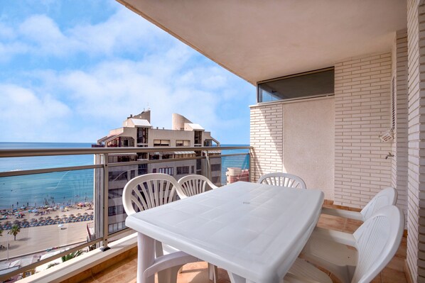 Un balcon avec une table et des chaises blanches, donnant sur une plage et un bâtiment à l’architecture moderne.