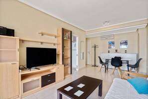 Un salon moderne doté d’un téléviseur à écran plat, de meubles en bois, d’un coin repas avec des chaises et d’une petite cuisine.