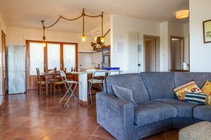 Een woonkamer met een bank, eettafel, stoelen en een keuken met een koelkast en oven.