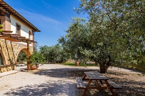 A building with stone arches, a wooden pergola, and a gravel path leading to a wooden picnic table.