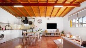 Una cuina moderna amb armaris blancs, una zona de menjador amb cadires de fusta, un televisor i una finestra gran.