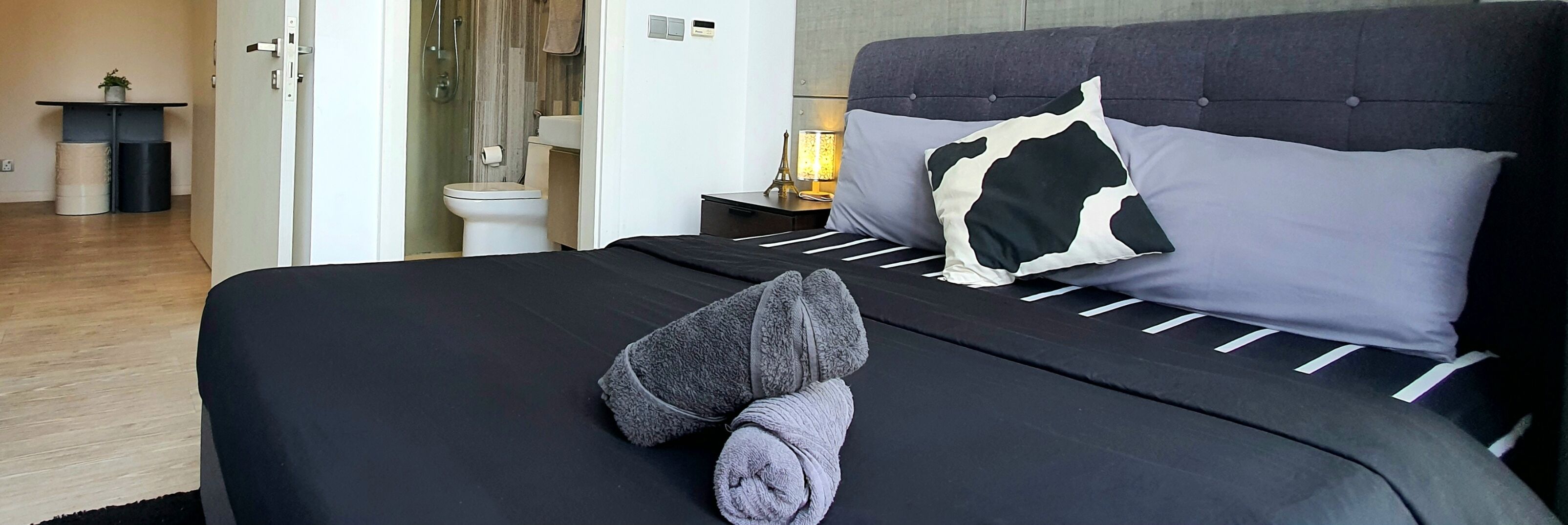 A neatly made bed with a black and white pillow, a pair of slippers, and a small bedside table with a lamp.