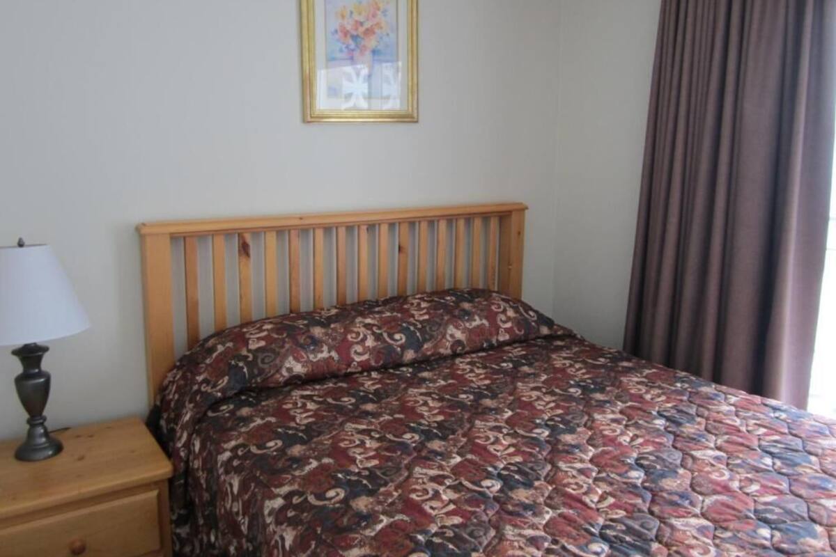 A bedroom with a wooden headboard, a patterned bedspread, a nightstand with a lamp, and a framed picture on the wall.