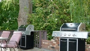 Two barbecue grills, a table with chairs, and a brick wall with greenery in the background. - Schell Motel (Vernon)