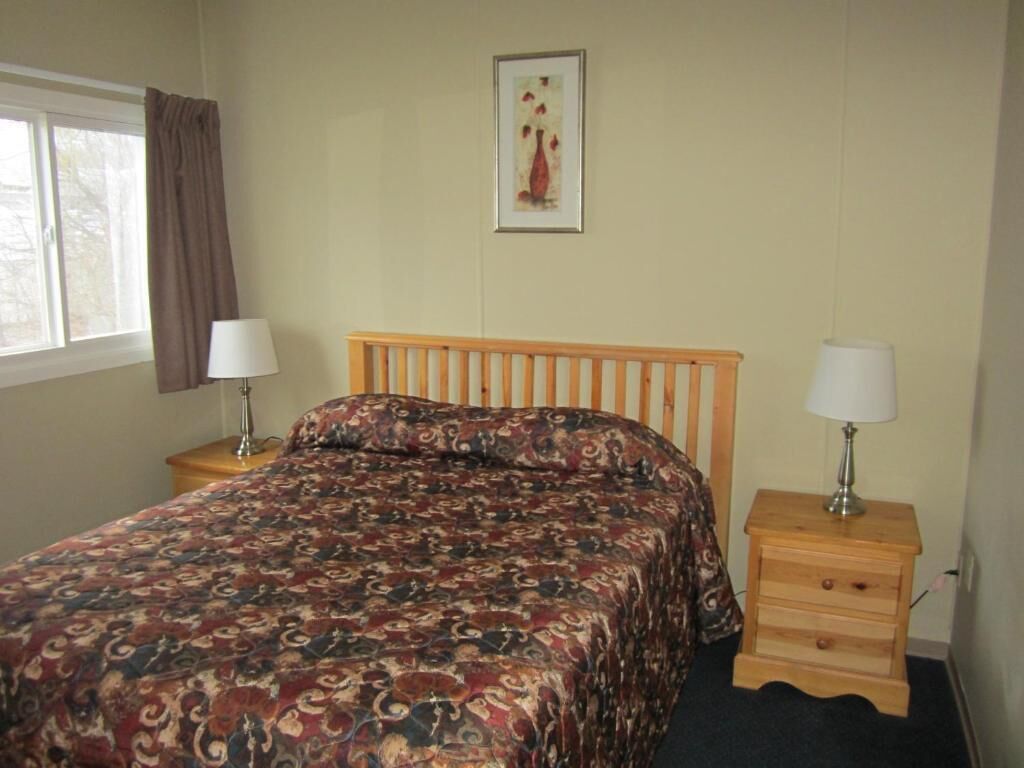 Un dormitorio con una cama de madera, dos mesitas de noche con lámparas, una ventana con cortinas y un cuadro en la pared.