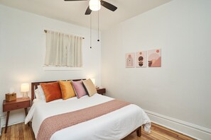 Una cama bien hecha con una colcha blanca y marrón, una mesita de noche de madera con una lámpara y un ventilador de techo.