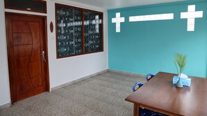 A room with a wooden door, a window with a patterned glass, a teal wall with two white crosses, a small table with a plant, and a blue chair.