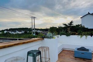 Una terraza en la azotea con cubierta de madera, un muro blanco con maceta y una mesa de vidrio transparente.