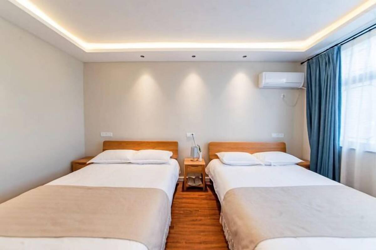 A hotel room with two single beds, a nightstand between them, an air conditioning unit on the wall, and a window with curtains.