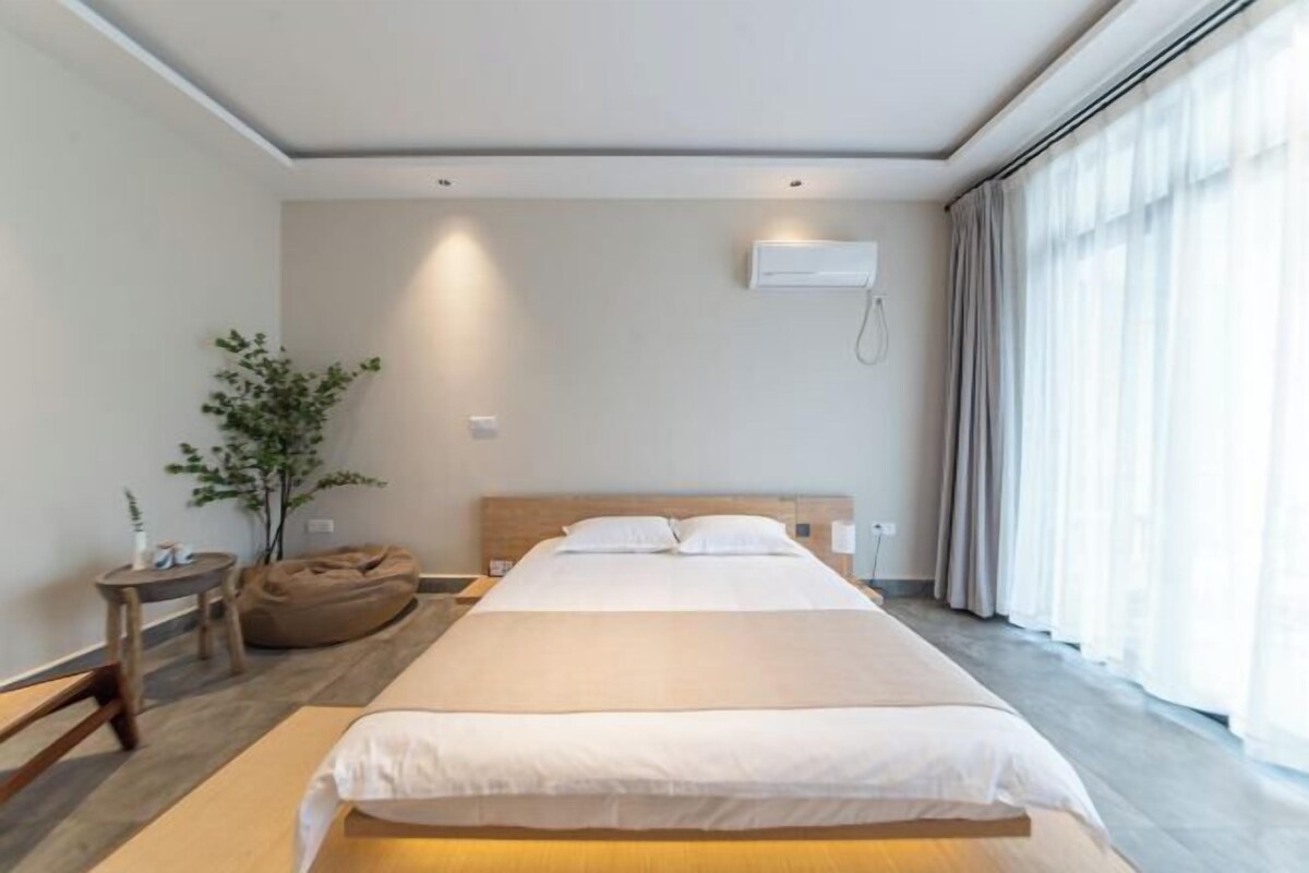A modern hotel room with a large bed, a bedside table, a plant, and a window with curtains.