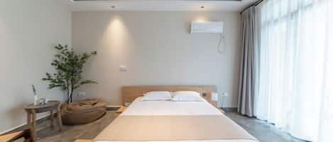 A modern hotel room with a large bed, a bedside table, a plant, and a window with curtains.