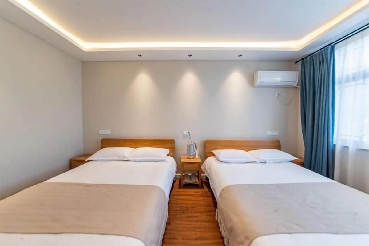 Ein Hotelzimmer mit zwei Betten, einem Nachttisch mit Lampe, einer Klimaanlage und einem Fenster mit Vorhängen.