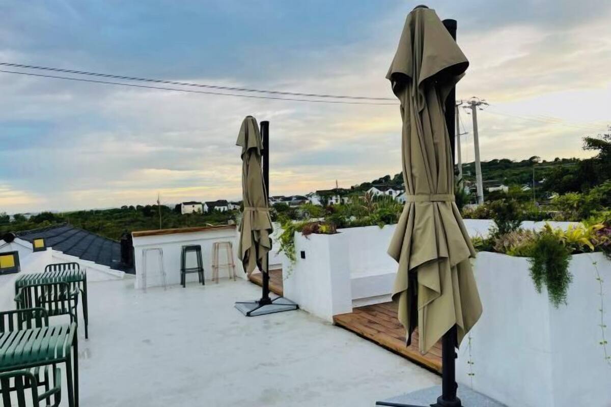 A rooftop terrace with outdoor seating and umbrellas, surrounded by greenery and houses in the background.