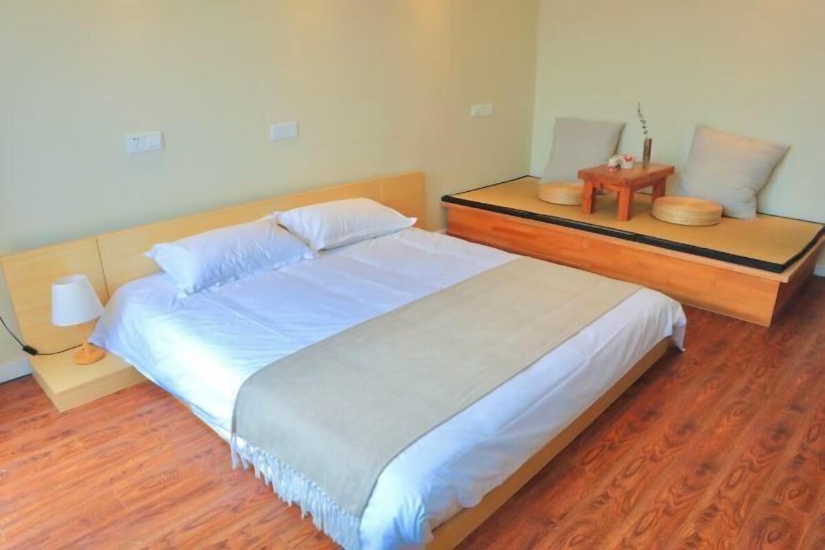 Quarto bem arrumado com cama, mesa de cabeceira, luminária e uma pequena mesa de madeira com almofadas.