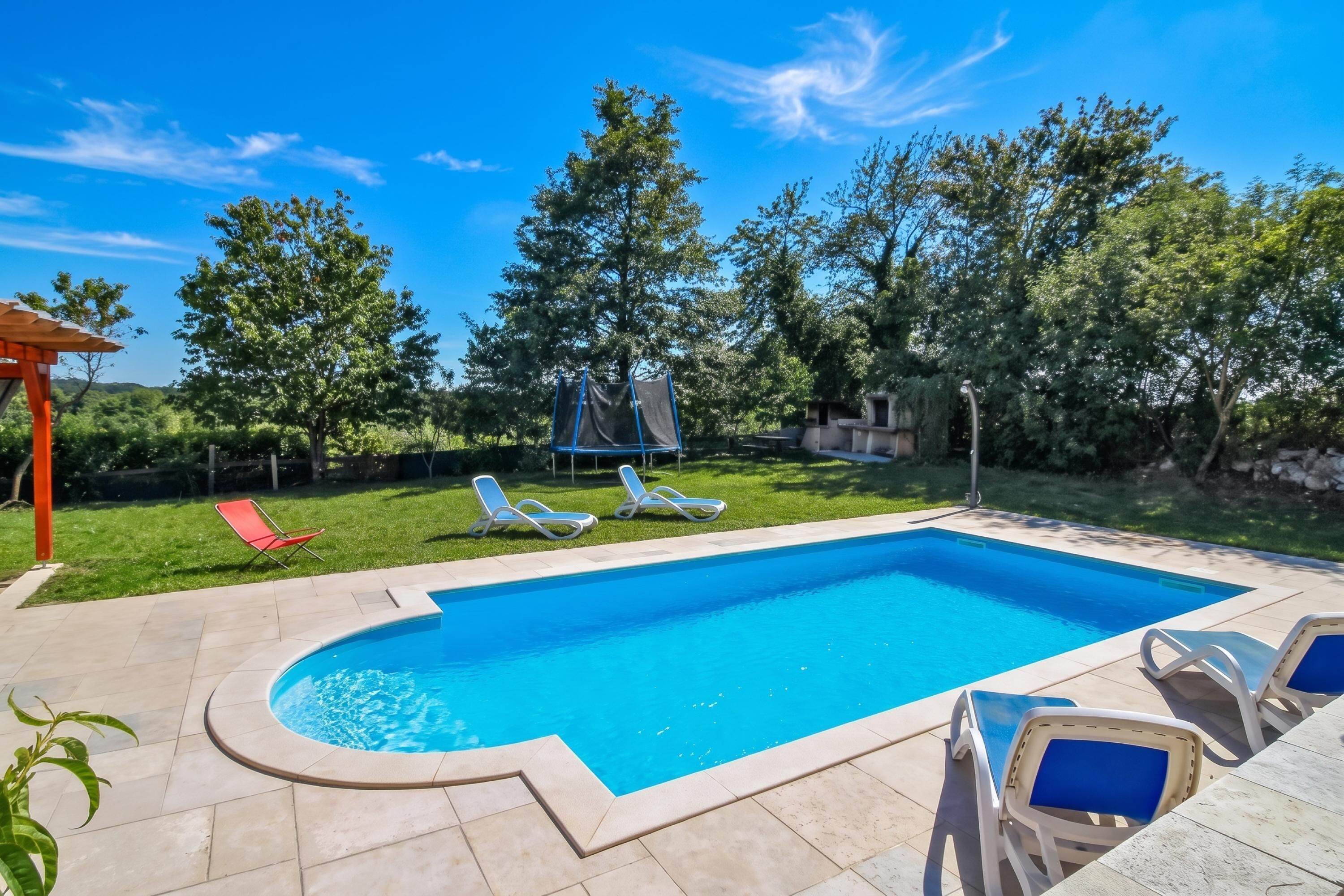 Une piscine rectangulaire avec une terrasse carrelée, entourée de chaises longues et d’une pelouse agrémentée d’arbres.