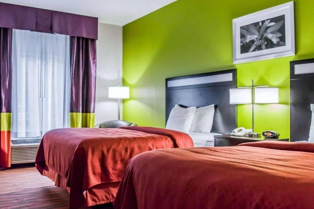 A hotel room with two beds, a large window with curtains, a green accent wall, and a framed picture.